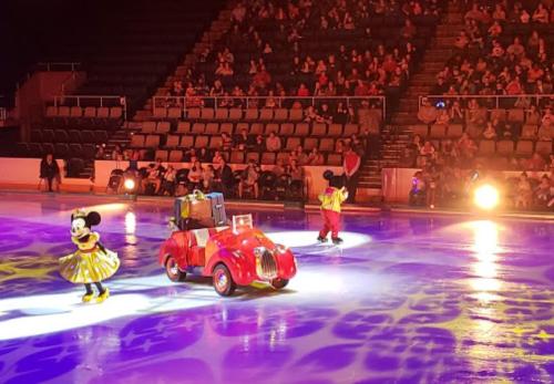 Minnie and Mickey Mouse perform on ice in front of a cheering audience, with a red car on the rink.