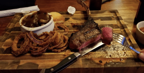 A wooden platter with a large steak, onion rings, a side of gravy, and a biscuit, with a fork and knife beside the steak.