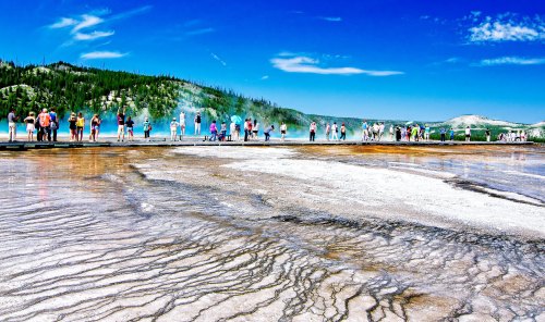 A vibrant landscape of Yellowstone National Park with visitors near a colorful hot spring and steam rising in the background.