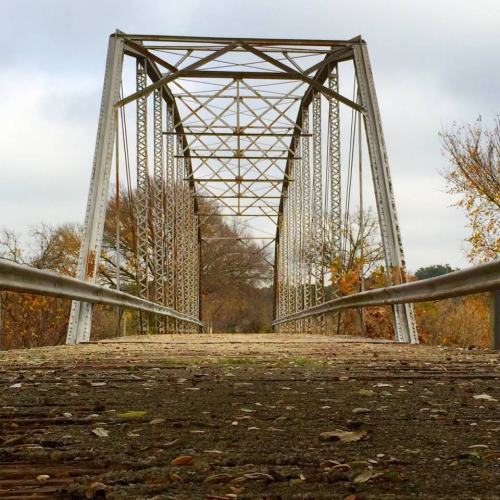 A weathered metal bridge stretches over a path, surrounded by trees with autumn foliage under a cloudy sky.