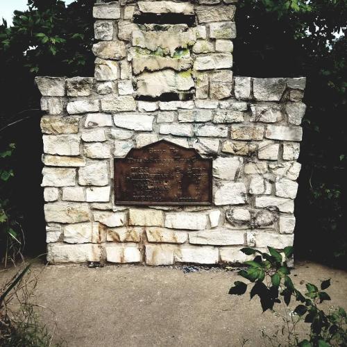 Stone monument with a weathered plaque, surrounded by greenery and concrete path.