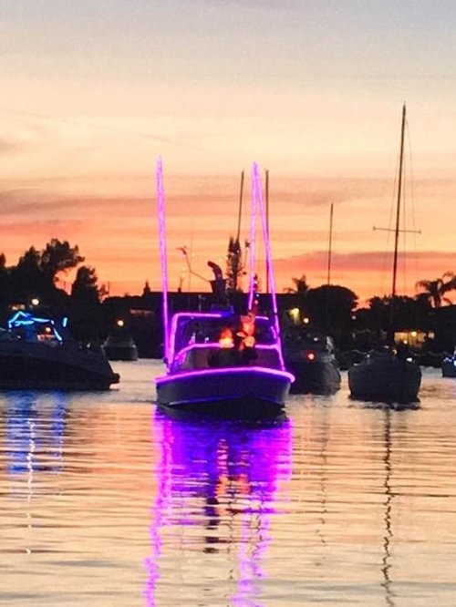A brightly lit boat with purple lights glides through calm waters at sunset, surrounded by other boats and a colorful sky.