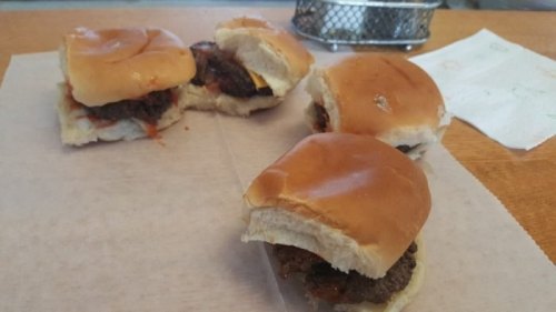 Four small sandwiches on a paper surface, featuring meat patties and soft, shiny buns.