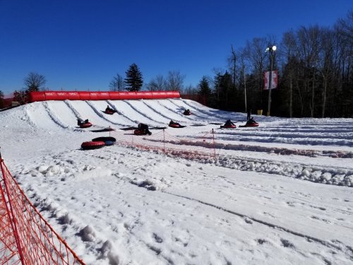 A snowy tubing hill with several people sliding down on inflatable tubes under a clear blue sky.