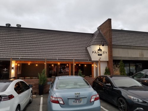 Exterior of a restaurant named "Pantry," featuring a sloped roof and outdoor seating area, with parked cars in front.