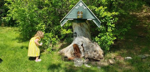 A young child in a yellow striped dress explores a whimsical birdhouse near a tree stump in a green garden.