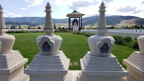 A serene landscape featuring ornate stupas in the foreground and a large Buddha statue in a pavilion against a mountainous backdrop.