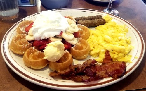 A plate of waffles topped with whipped cream, strawberries, bananas, scrambled eggs, sausage, and bacon.