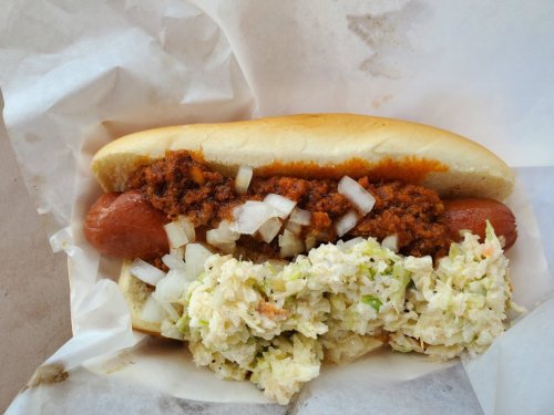 A hot dog topped with chili and onions, served alongside a portion of creamy coleslaw in a paper wrapper.