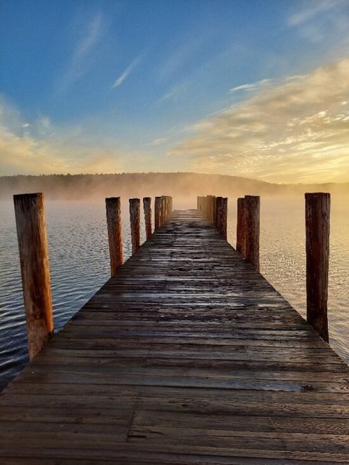 A wooden pier extends into a misty lake at sunrise, surrounded by calm water and a colorful sky.