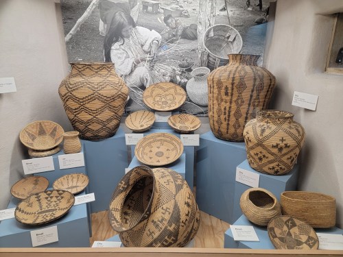 Display of intricately woven baskets and pottery, showcasing various patterns and designs, with a background image of weaving.