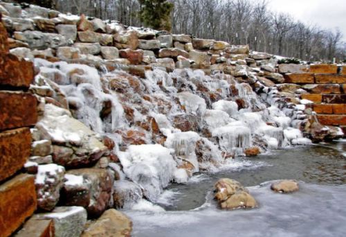 A frozen waterfall cascading over stone steps, surrounded by icy rocks and bare trees in a winter landscape.