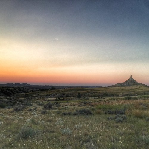 A serene landscape at sunset, featuring a distant rock formation and rolling hills under a colorful sky.