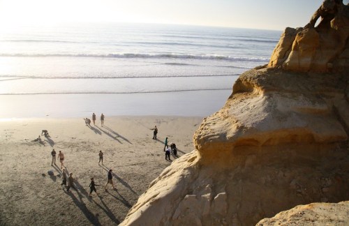 People walking and playing on a sandy beach near cliffs, with gentle waves lapping at the shore under a bright sky.