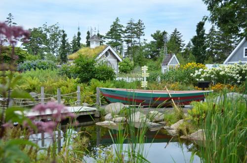 A colorful boat rests by a serene pond, surrounded by lush greenery and blooming flowers, with quaint buildings in the background.