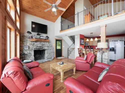 Cozy living room with red leather sofas, stone fireplace, wooden ceiling, and open kitchen area. Bright and inviting space.
