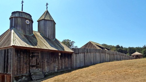 Historic wooden church with two domed towers, surrounded by a wooden fence and grassy landscape under a clear blue sky.
