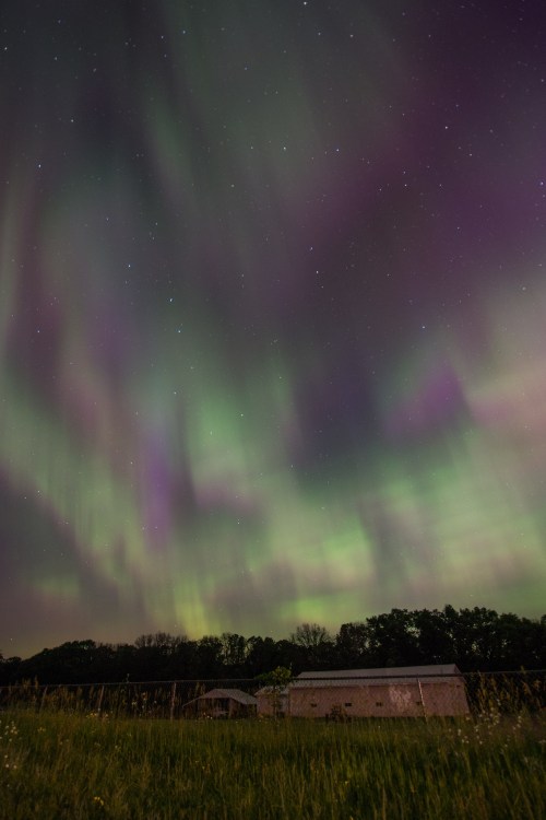 A vibrant display of the northern lights illuminates the night sky above a rural landscape and a small building.
