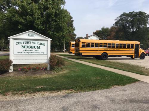 Sign for Century Village Museum with a yellow school bus parked nearby on a sunny day.