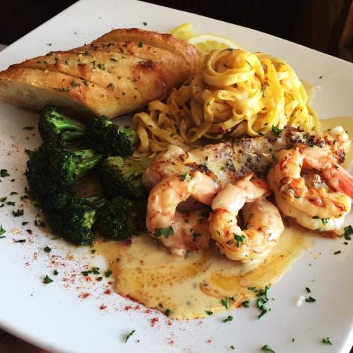 A plate of shrimp with creamy sauce, fettuccine, broccoli, and garlic bread, garnished with herbs and lemon.