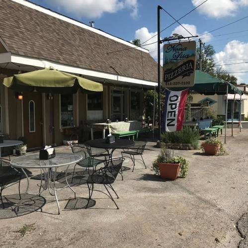 A quaint pizzeria with outdoor seating, featuring a sign that says "Open" and a sunny blue sky above.