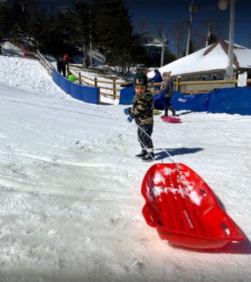 A child in a camouflage jacket pulls a red sled on a snowy hill, with other people sledding in the background.