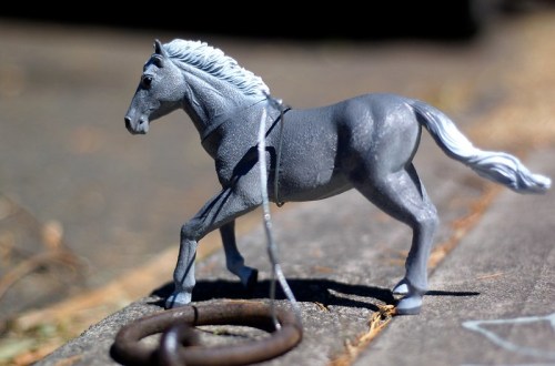 A gray toy horse stands on a wooden surface next to a metal ring, with a blurred background.