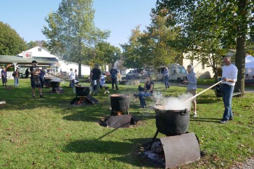 People cooking outdoors in large pots over open flames, surrounded by trees and a clear blue sky.
