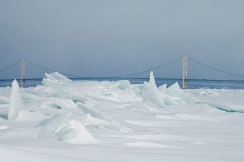 A snowy landscape with ice formations in the foreground and a bridge in the background under a cloudy sky.