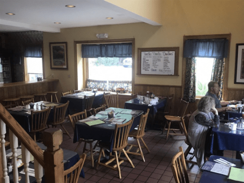 A cozy restaurant interior with wooden tables, blue tablecloths, and a few diners enjoying their meals.