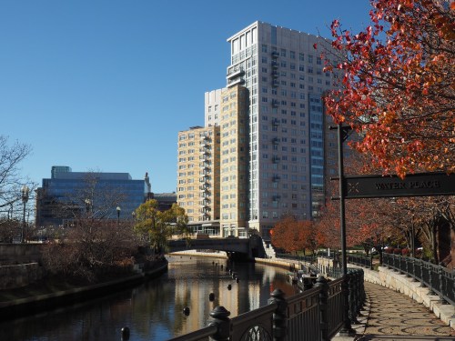 A scenic view of a river with buildings and autumn foliage along the walkway on a clear day.