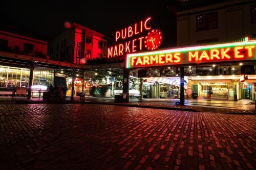Neon signs illuminate a brick-paved market at night, showcasing "Public Market" and "Farmers Market."