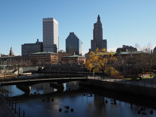 City skyline with modern and historic buildings, a river, and autumn trees under a clear blue sky.