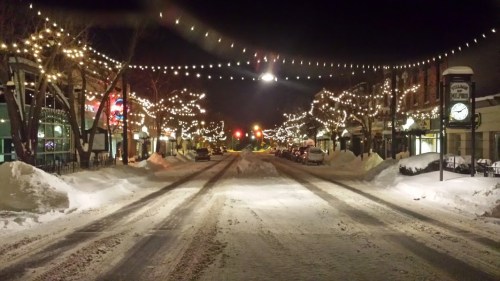 Snow-covered street at night, lined with festive lights and empty shops, creating a serene winter atmosphere.