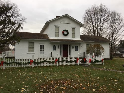 A white house with a wreath on the door, surrounded by a picket fence decorated with red bows, set in a grassy area.