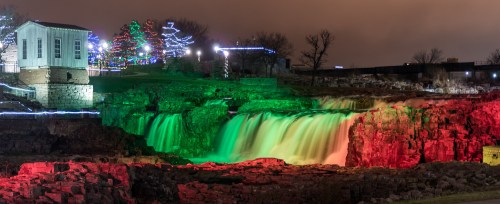 A waterfall illuminated in green and red lights, with a building and festive trees in the background at night.