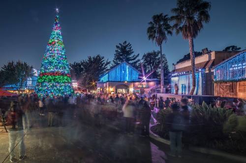 A festive scene with a brightly lit Christmas tree and people enjoying holiday decorations at night.