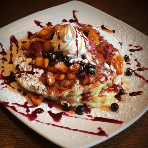 A stack of fluffy pancakes topped with fresh fruit, whipped cream, and a drizzle of berry sauce on a white plate.