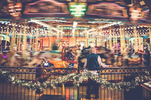 A blurred carousel with colorful lights, featuring a couple standing by a festive railing, enjoying the scene.