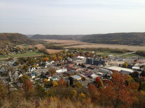 Aerial view of a small town surrounded by rolling hills and fields, showcasing autumn foliage and a clear sky.