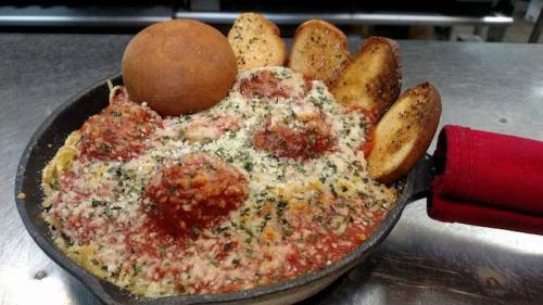 A skillet filled with meatballs in marinara sauce, topped with cheese and served with toasted bread and a roll.