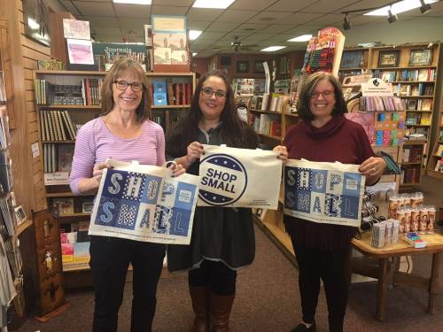 Three women hold "Shop Small" tote bags in a cozy bookstore filled with books and products.