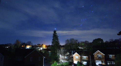 A night sky filled with stars over a suburban neighborhood, featuring a tall tree and illuminated houses.