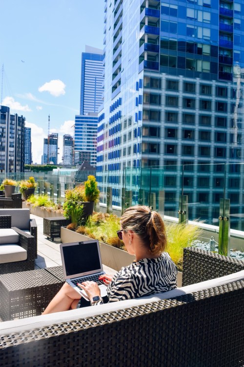 A woman works on a laptop on a rooftop terrace surrounded by modern buildings and greenery.