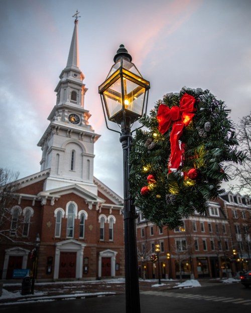 A festive wreath with a bow hangs on a lamp post, with a church steeple in the background under a cloudy sky.