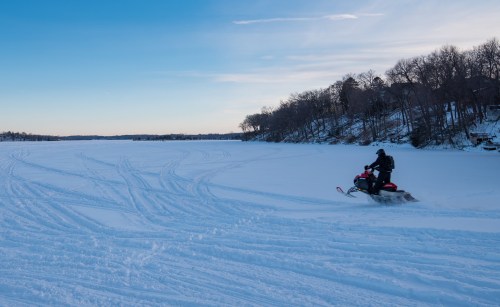A snowmobiler rides across a snowy lake under a clear blue sky, with trees lining the shore in the background.