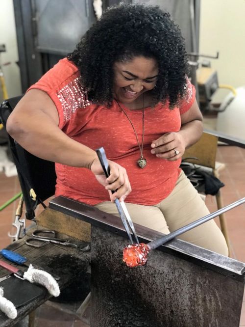 A woman with curly hair smiles while shaping molten glass in a glassblowing studio.