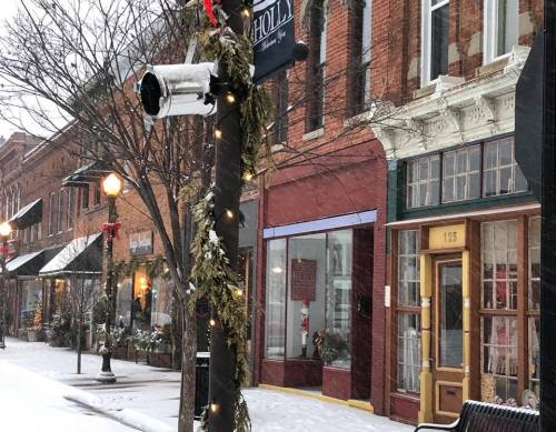 Snowy street scene with festive decorations, shops, and a lamppost adorned with greenery and lights.