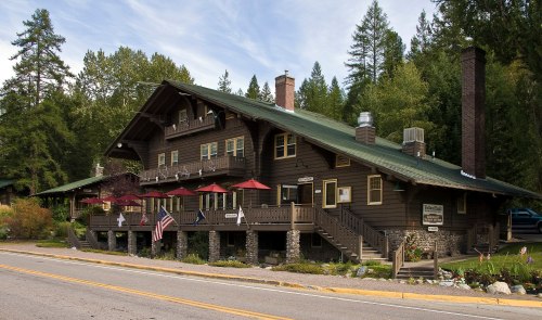 A large, rustic wooden lodge with a green roof, surrounded by trees and featuring a porch with red umbrellas.