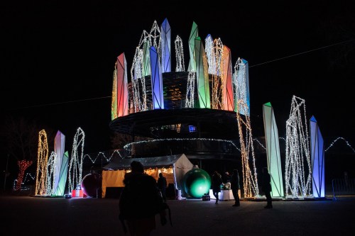A colorful illuminated structure with crystal-like shapes, surrounded by festive lights and people at night.
